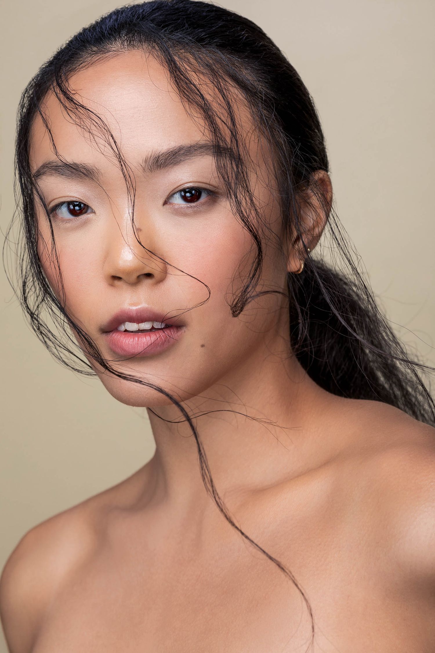 Young Asian woman with wet hair and natural makeup poses for a beauty portrait against a neutral beige background.