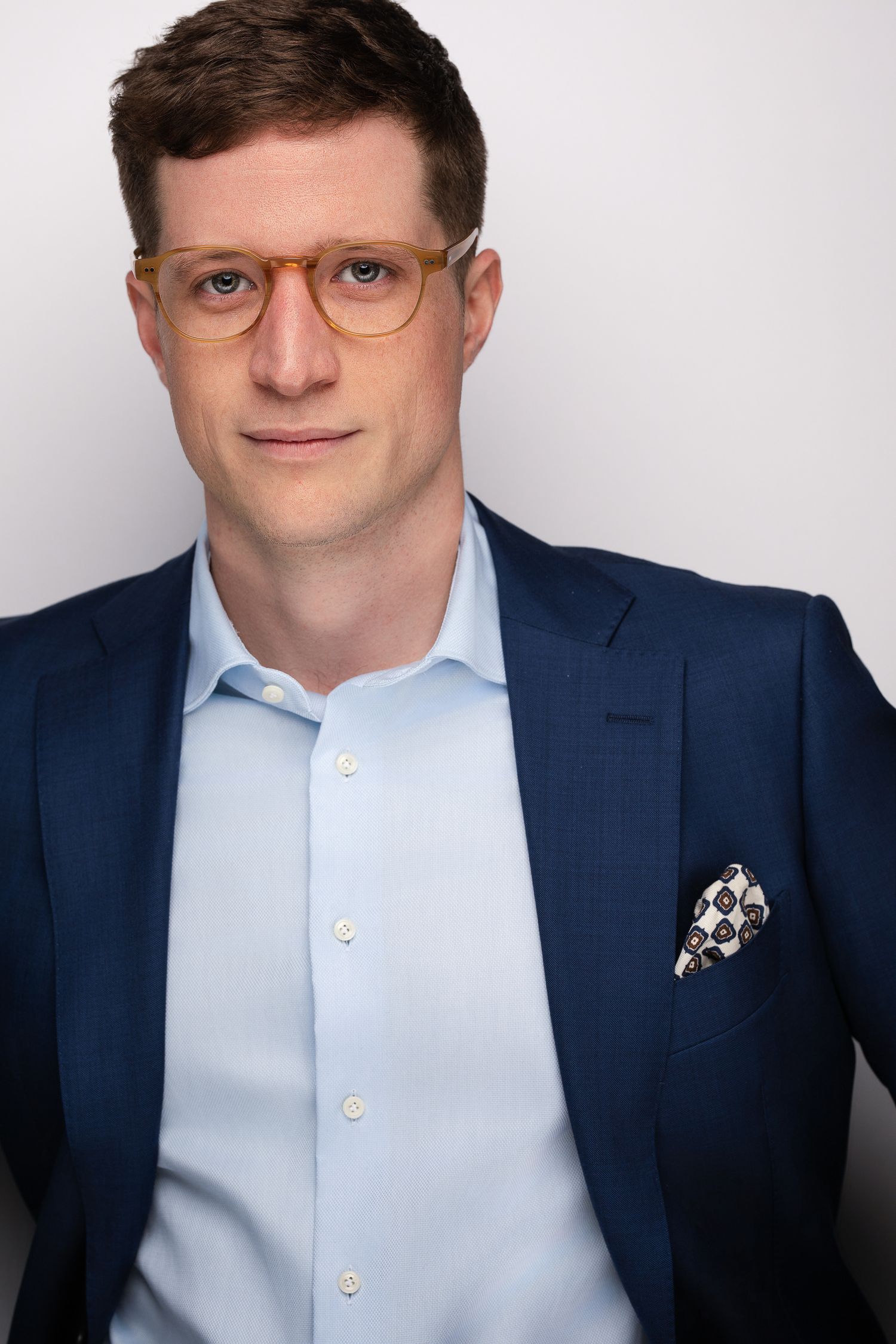 Man in navy blazer and light blue shirt with patterned pocket square and amber glasses poses for professional headshot.