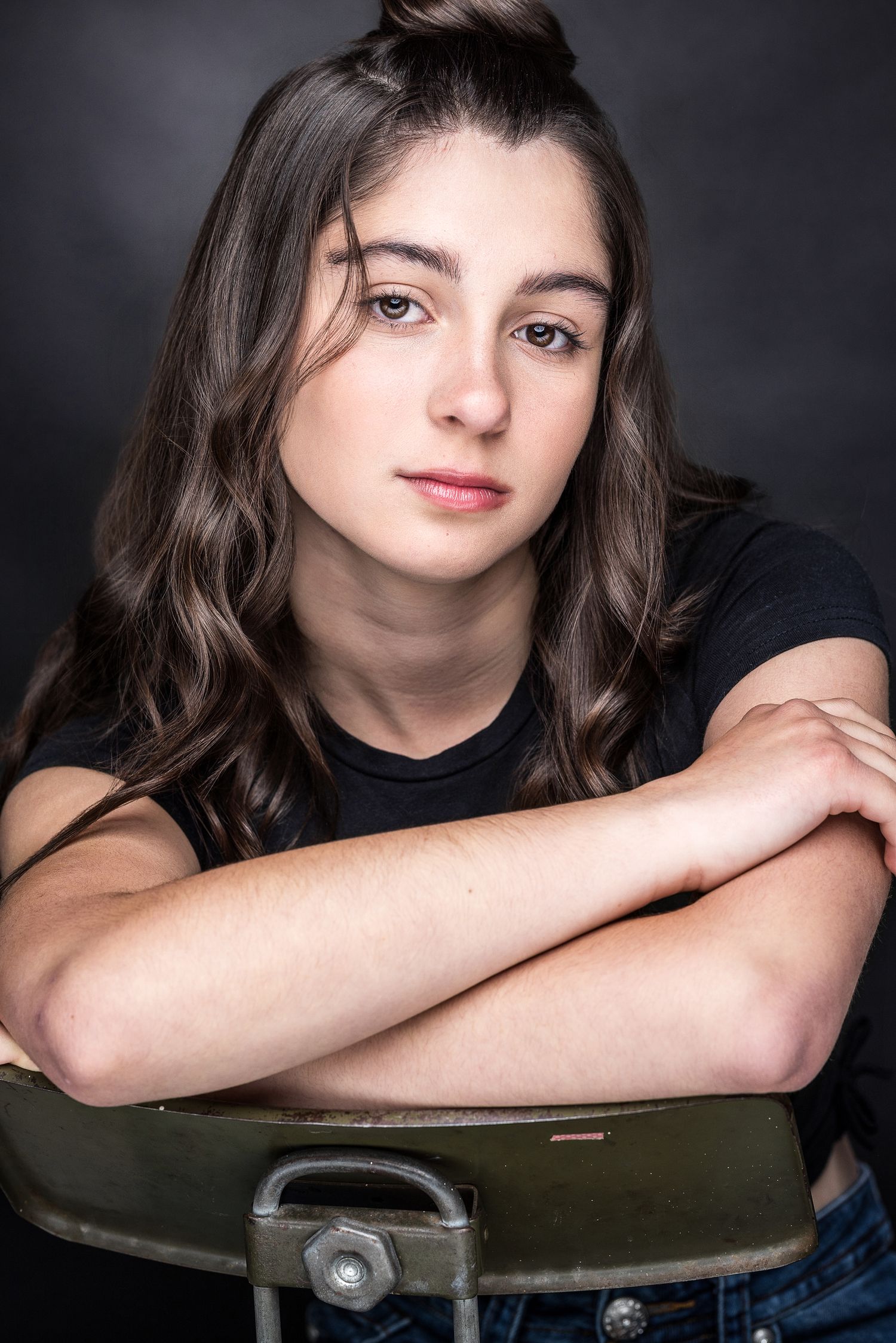 Young woman with wavy brown hair poses with arms crossed on chair back against dark studio background.