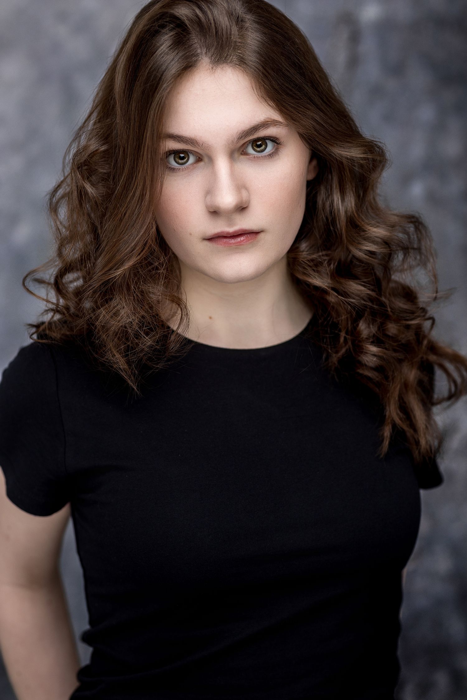 Young woman with curly brown hair wearing a black t-shirt poses for a professional headshot photo.