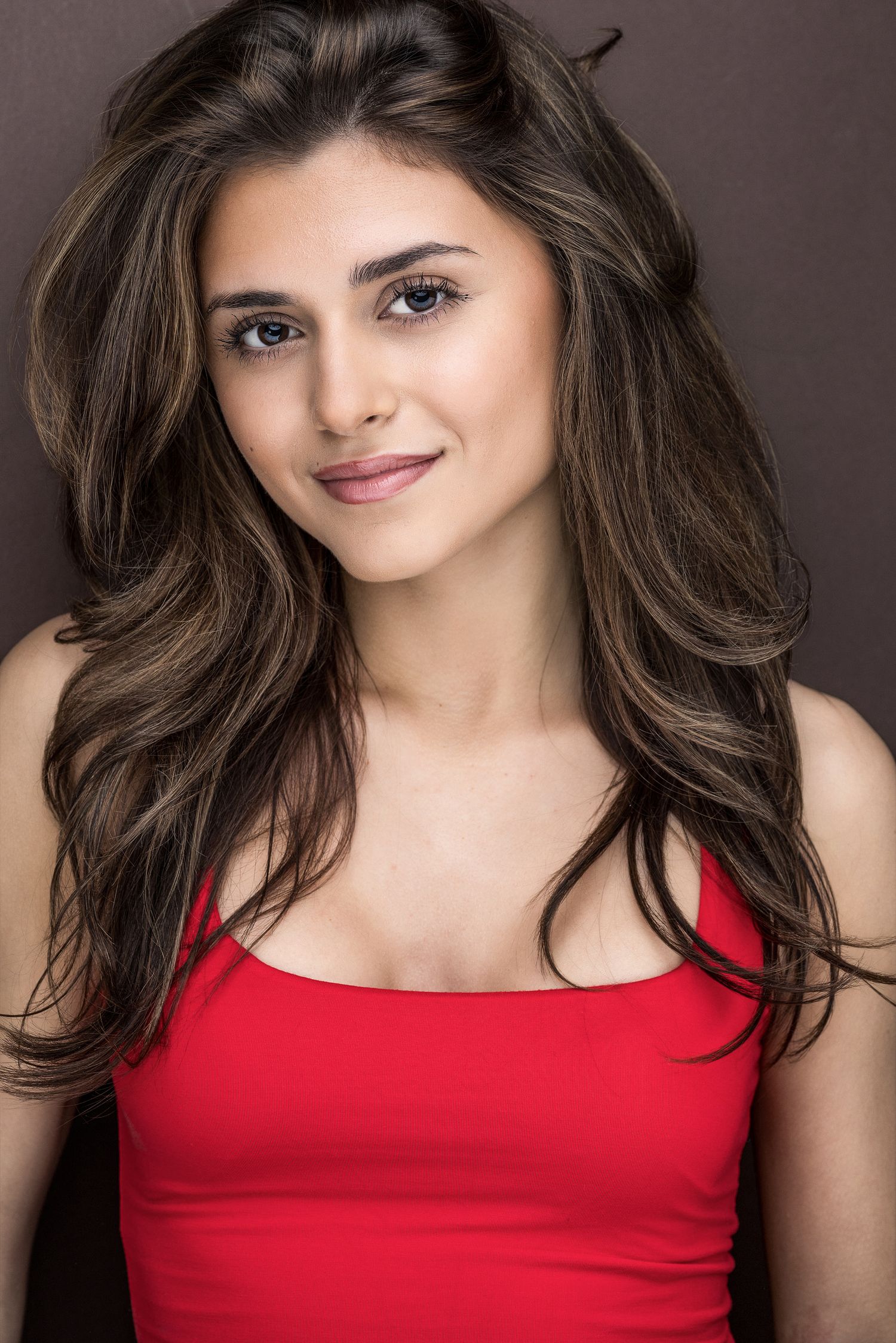 Young brunette woman in a red tank top smiling softly against a dark brown studio background.