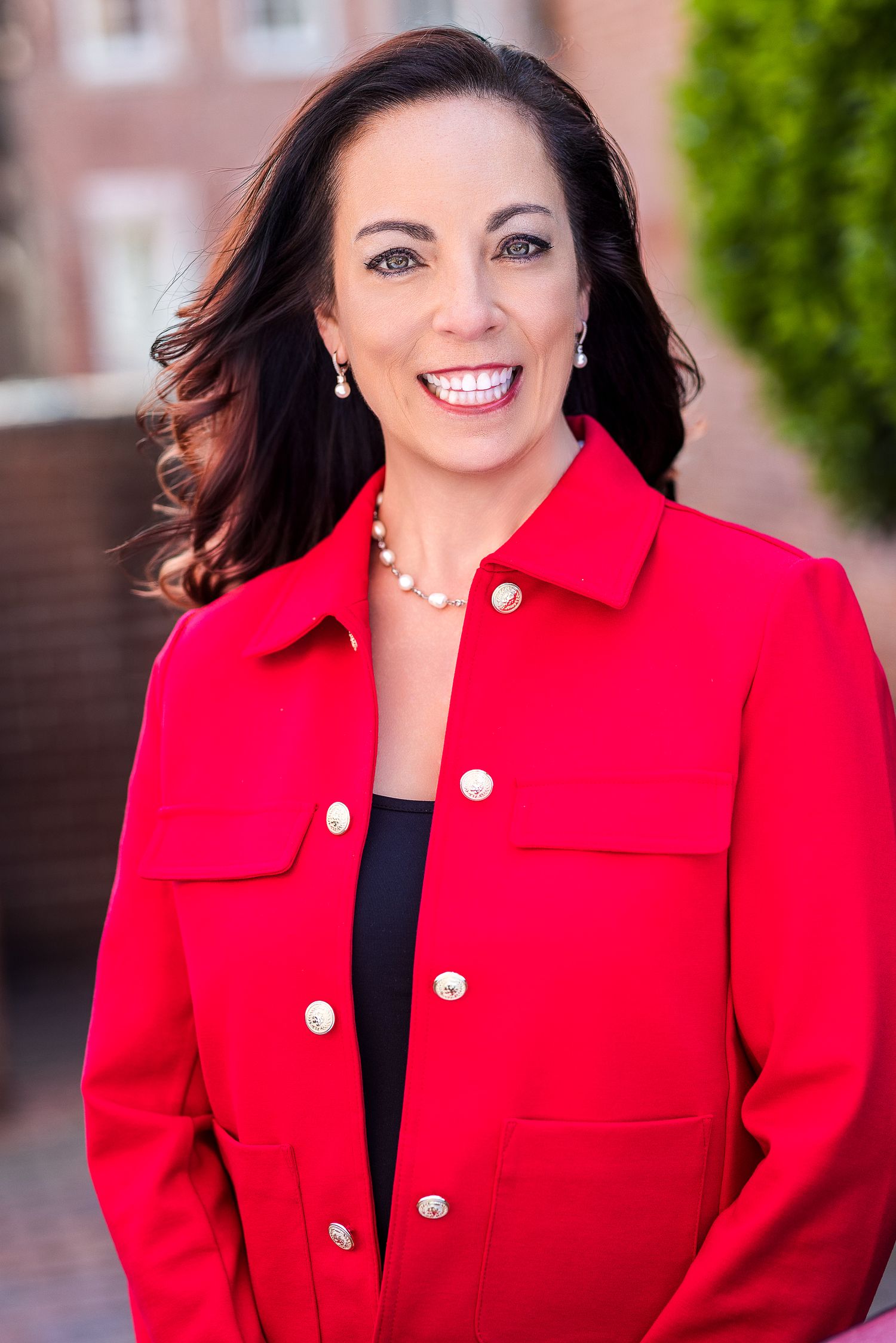 Professional woman in red blazer with pearl jewelry smiling outdoors in a polished business portrait.