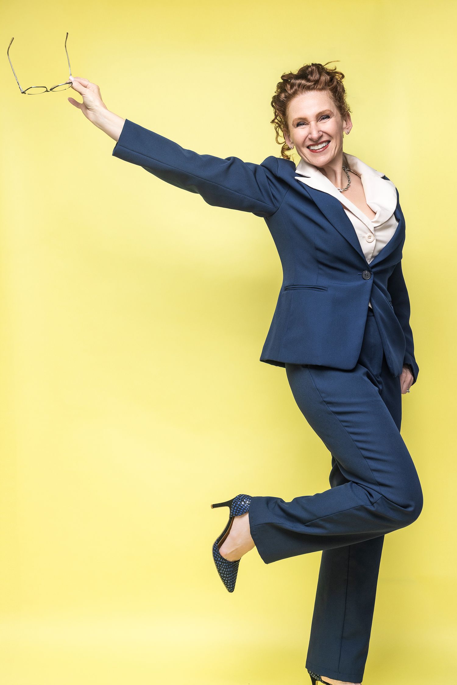 Cheerful businesswoman in navy suit jumping energetically against yellow background while holding glasses.