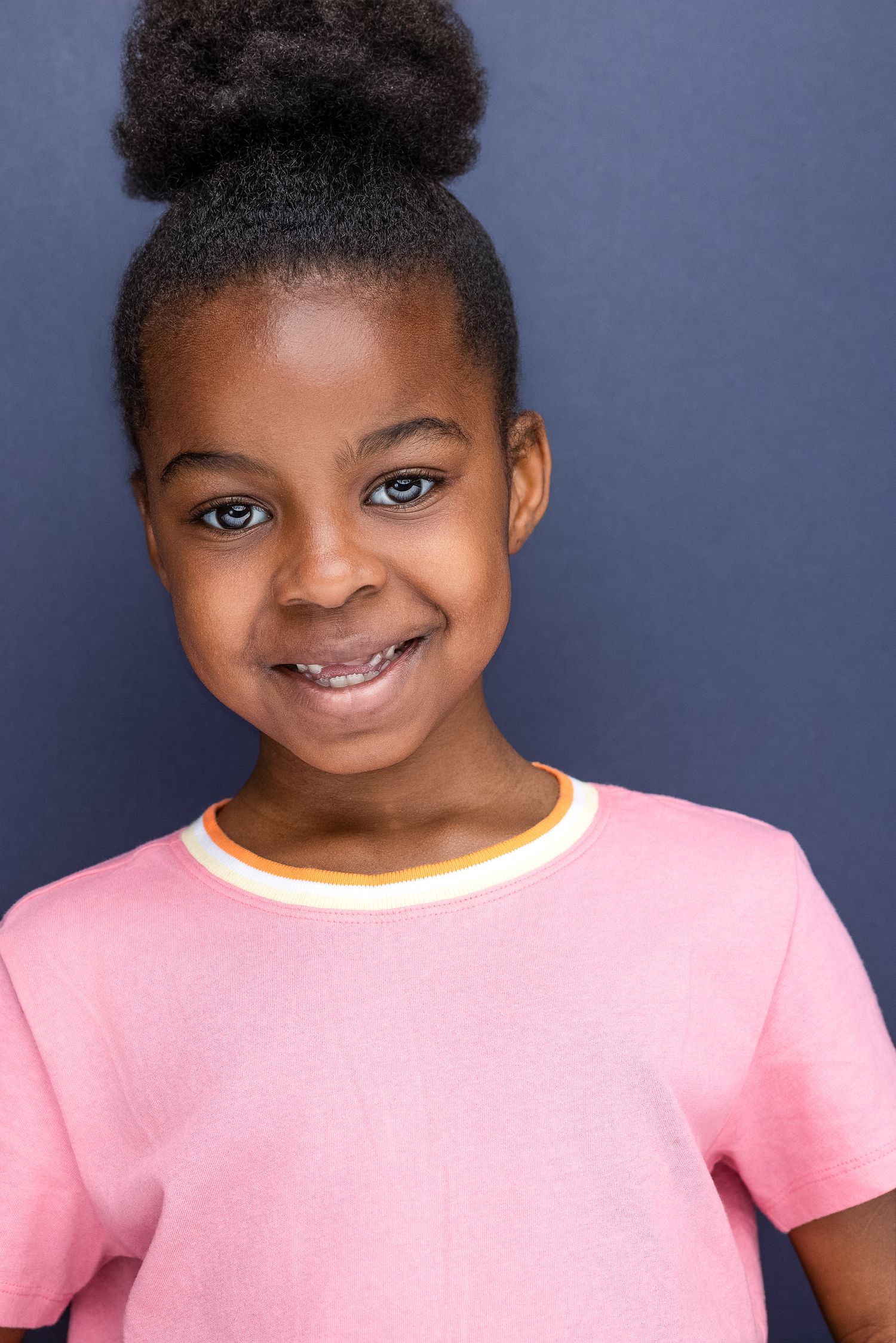 Young girl smiling in pink t-shirt with hair in bun against blue background.