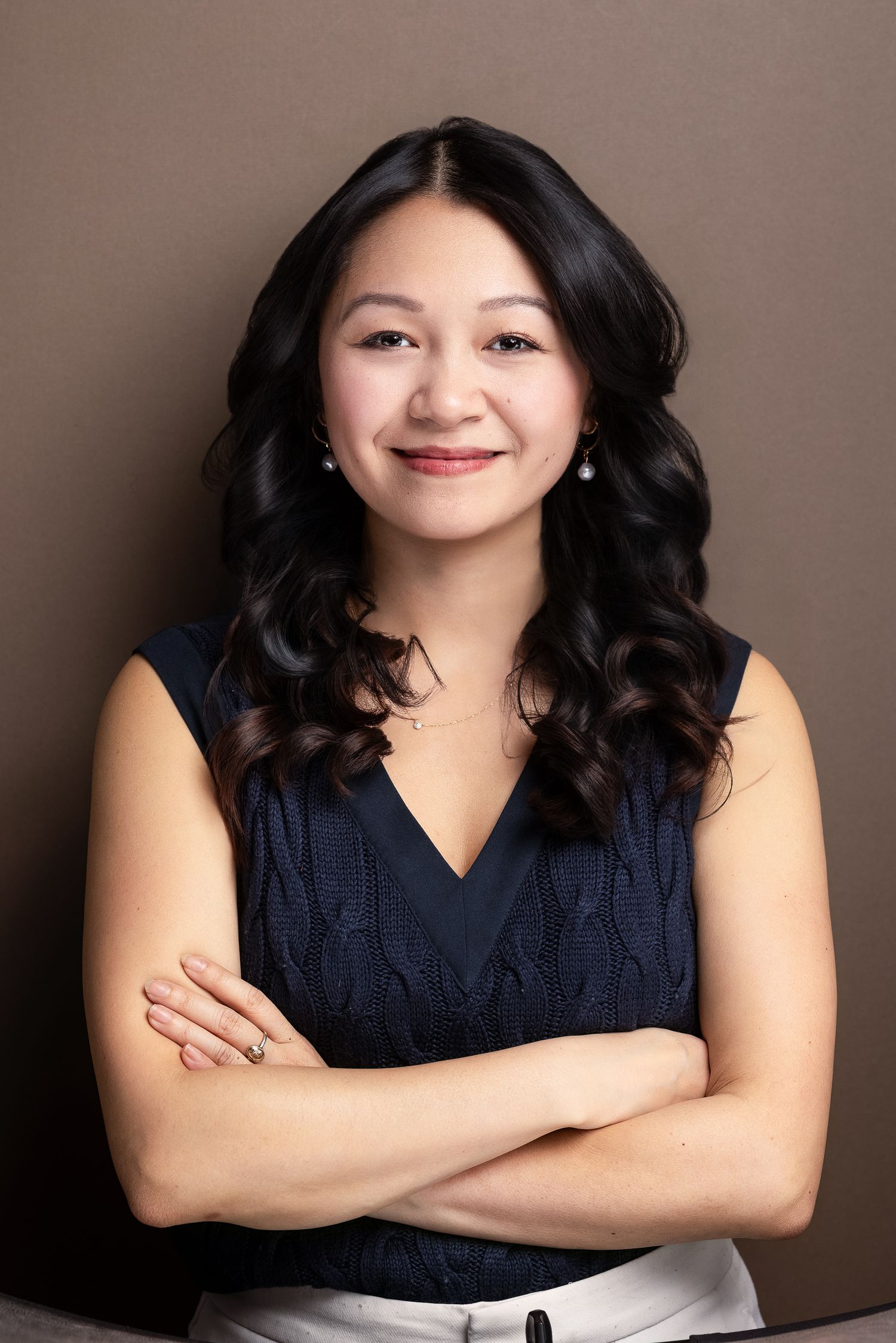 Professional headshot of a smiling young woman with curly black hair, wearing a navy sleeveless top, arms crossed.