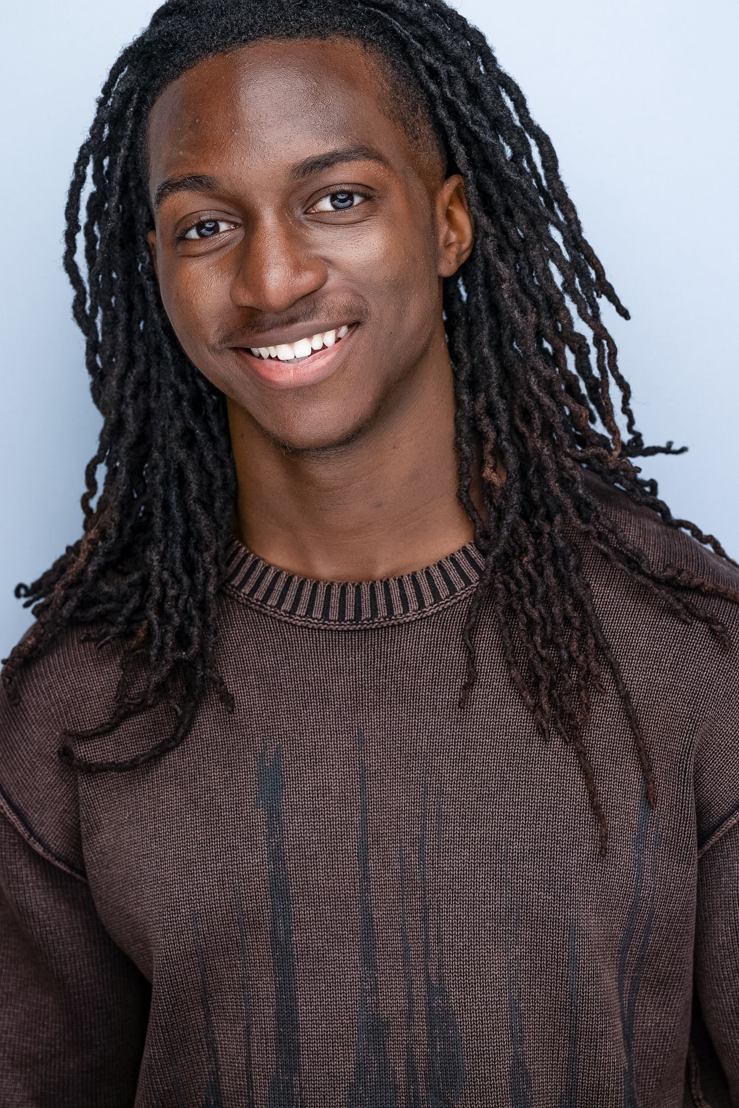 Young Black man with long dreadlocks wearing brown sweater smiles for professional headshot on blue background.