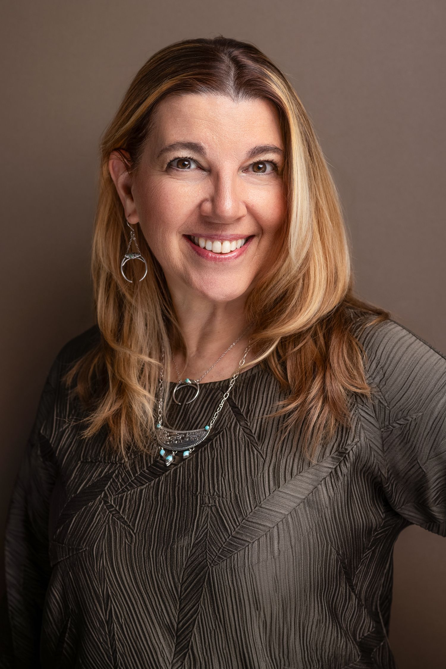 Smiling woman with blonde highlights wearing a gray patterned blouse and layered necklace against a brown background.