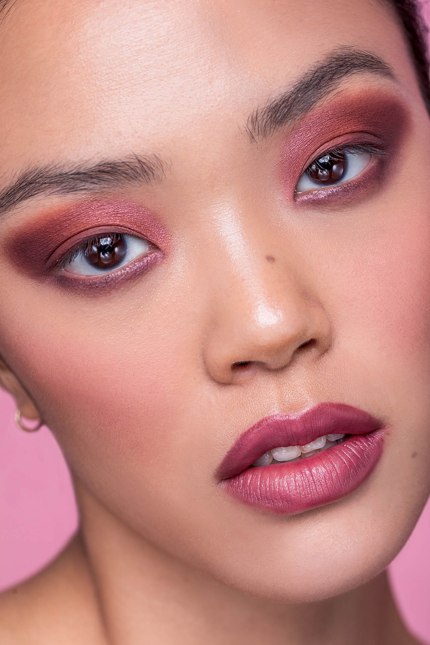 Close-up beauty portrait of a woman wearing bold pink-red smoky eye makeup and matching glossy lips on pink background.
