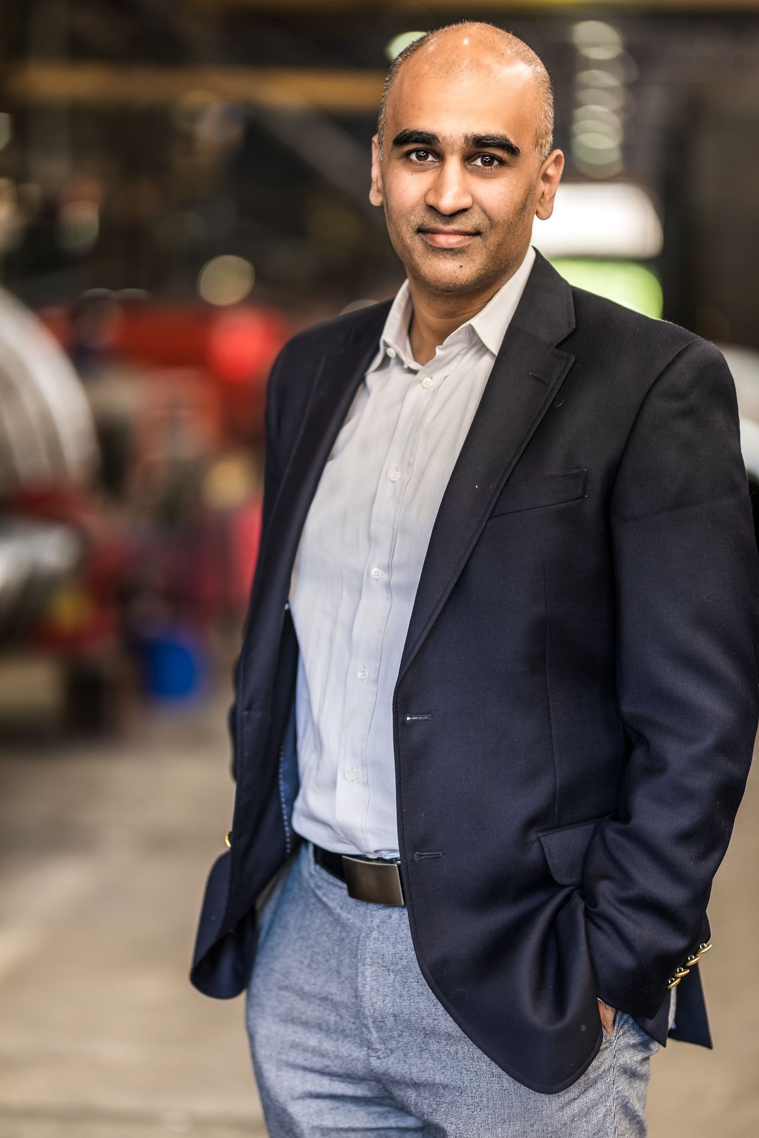 Professional headshot of a bald man in a navy blazer and gray trousers standing in an industrial setting.