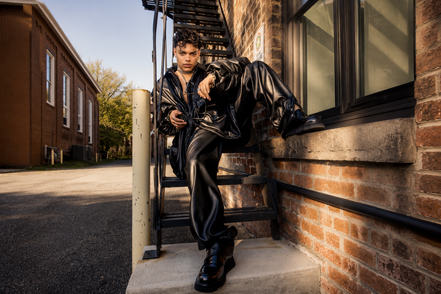 Young man in black leather outfit sits confidently on outdoor stairs in a stylish fashion portrait.