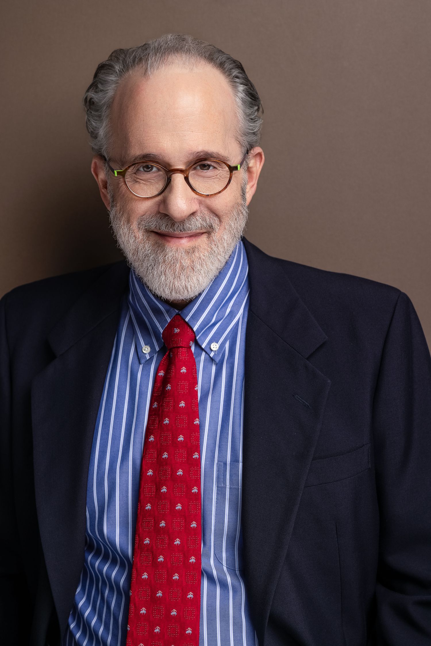 Professional headshot of a bearded man with glasses wearing a blue striped shirt, red tie, and dark blazer.