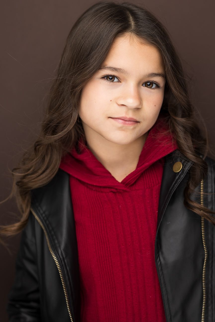 Young girl with long dark hair wearing a red hoodie and black leather jacket, posing for a portrait photo.