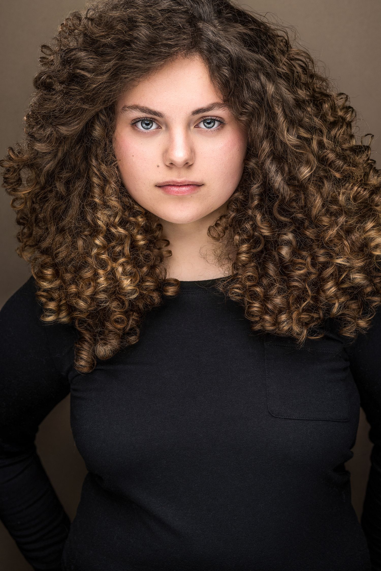 Young woman with voluminous curly brown hair and blue eyes wearing a black top, professional headshot photo.