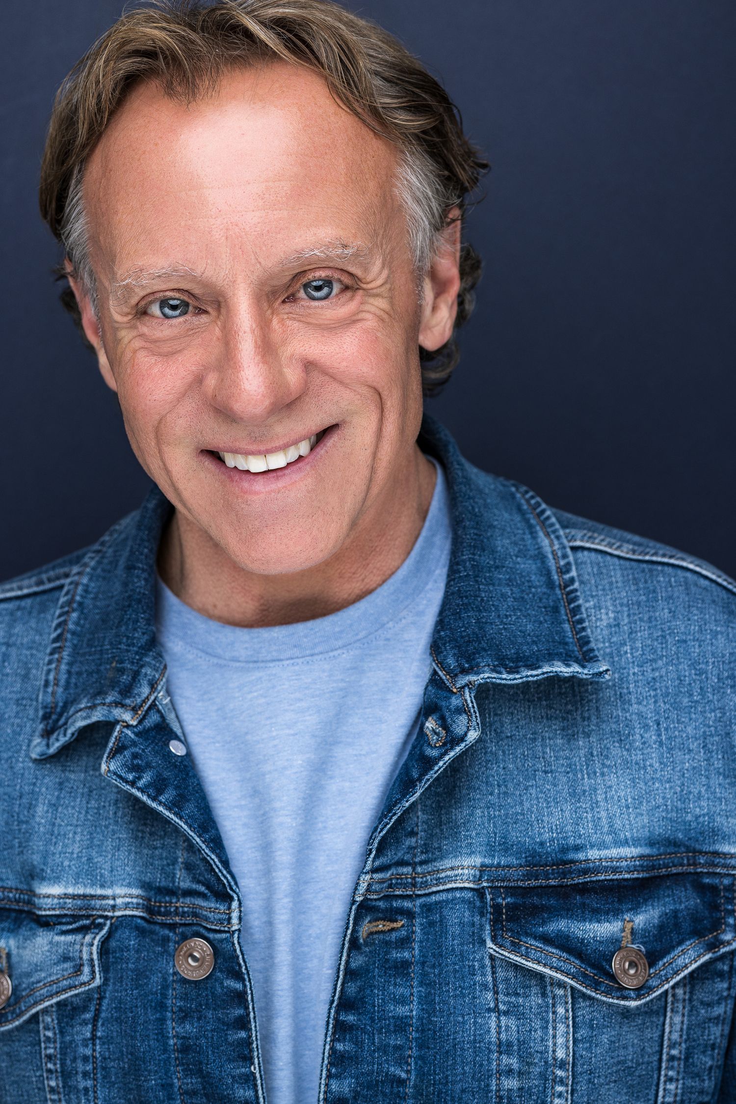 Smiling middle-aged man with blue eyes wearing a denim jacket over a light blue shirt against a dark background.
