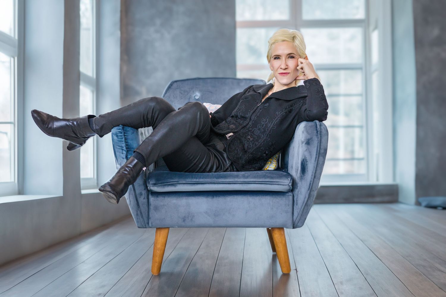 Blonde woman in black outfit sitting relaxed in a blue velvet armchair, legs up, in a bright modern room.