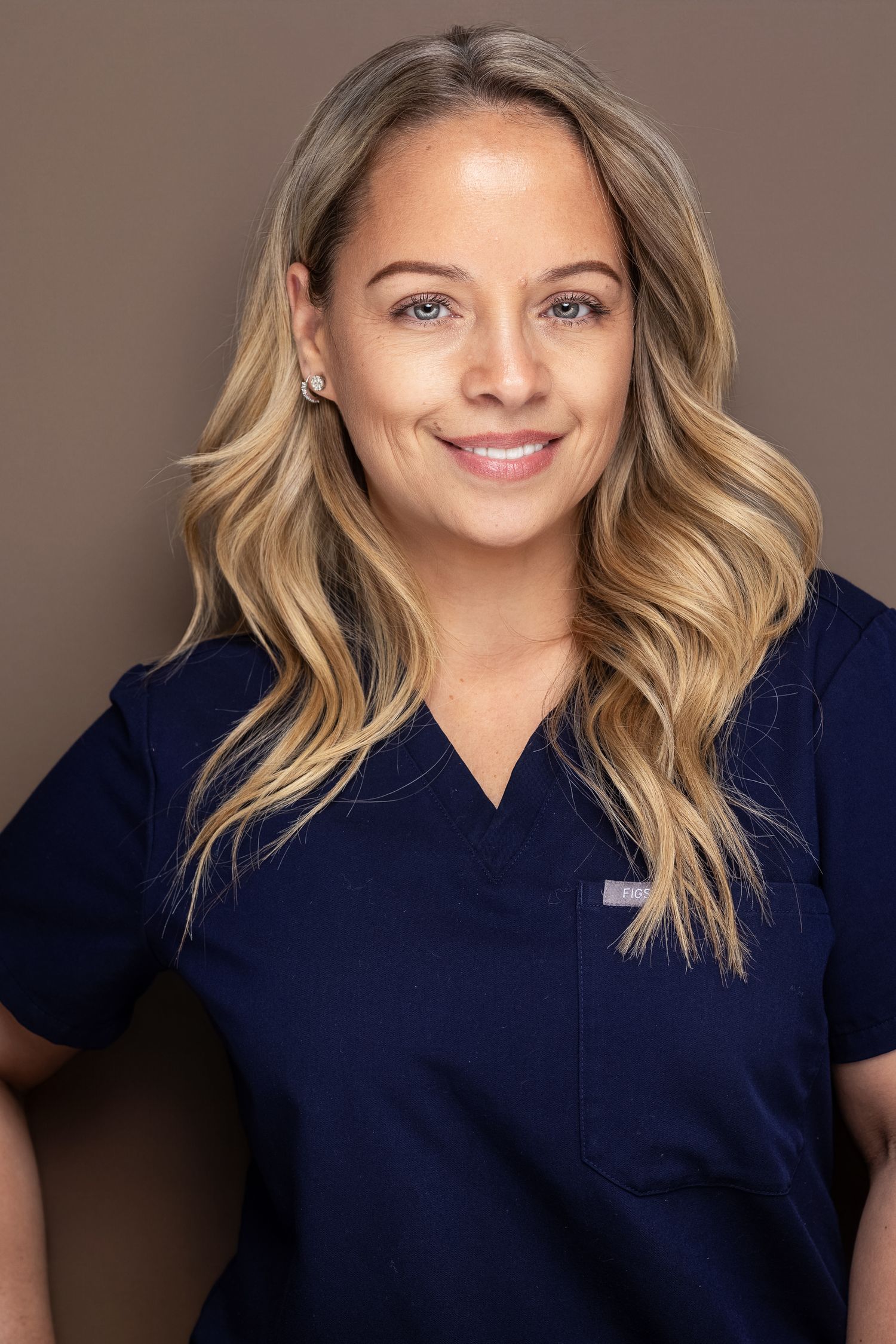 Blonde woman in navy blue medical scrubs smiling against brown background.