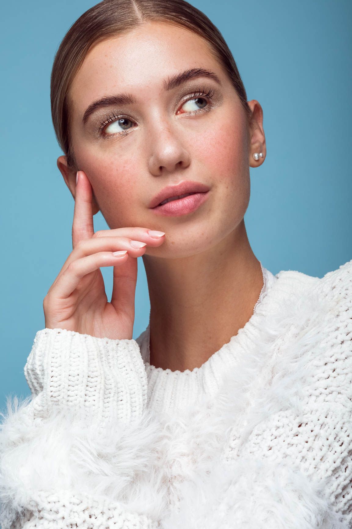 Young woman in white fluffy sweater with natural makeup and pearl earring poses thoughtfully against a blue background.