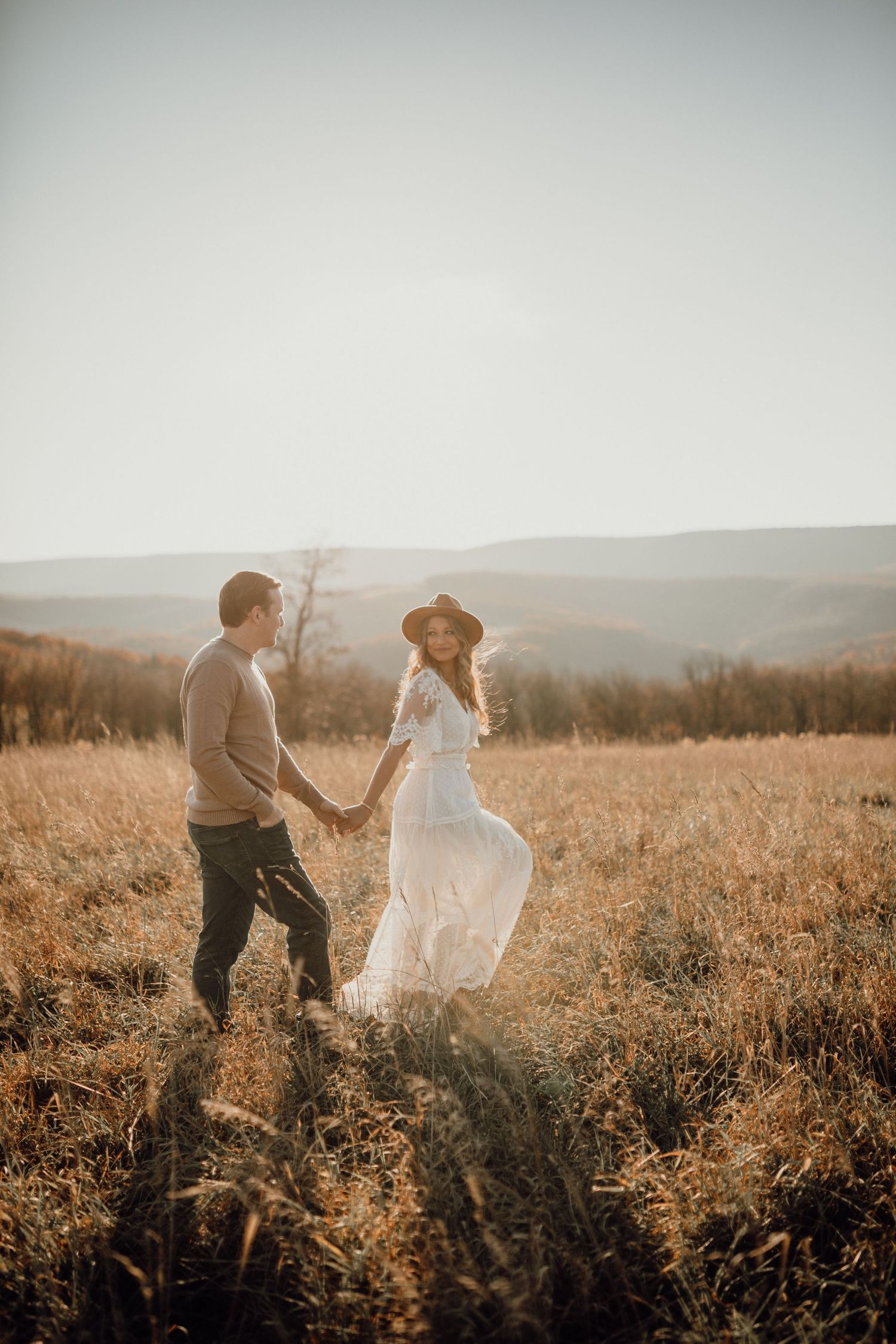 Couple holds hands and walks through a golden autumn field with rolling hills behind them, the woman in a white dress and hat.