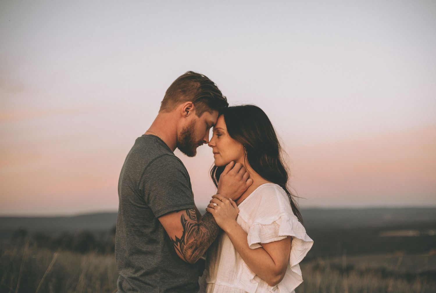 Couple touching foreheads in a tender moment at dusk, the tattooed man cradling his partner's face with a pastel sky behind.