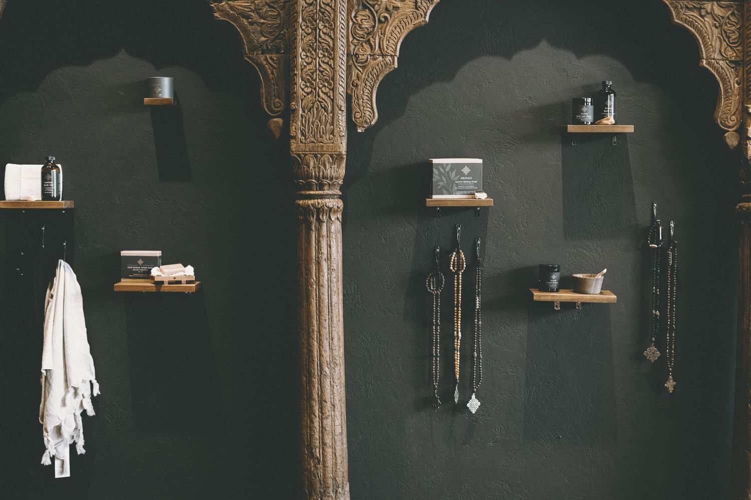 Dark charcoal wall with small wooden shelves holding skincare items, hanging jewelry and a white towel beside a carved wooden pillar.