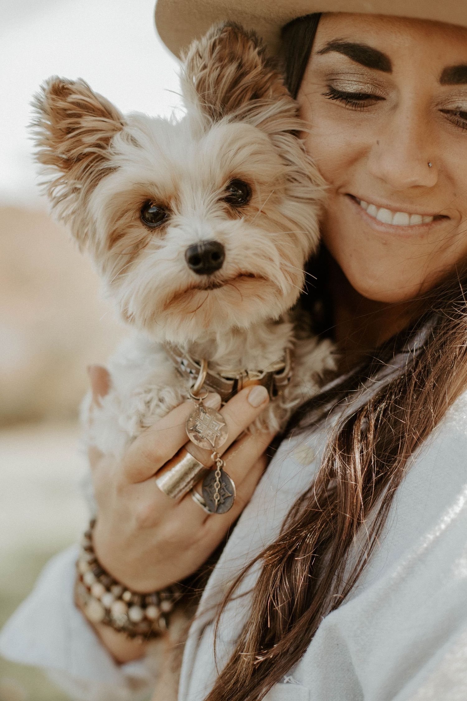 Smiling woman in a tan hat holding a small Yorkshire terrier wearing a collar with charms outdoors.