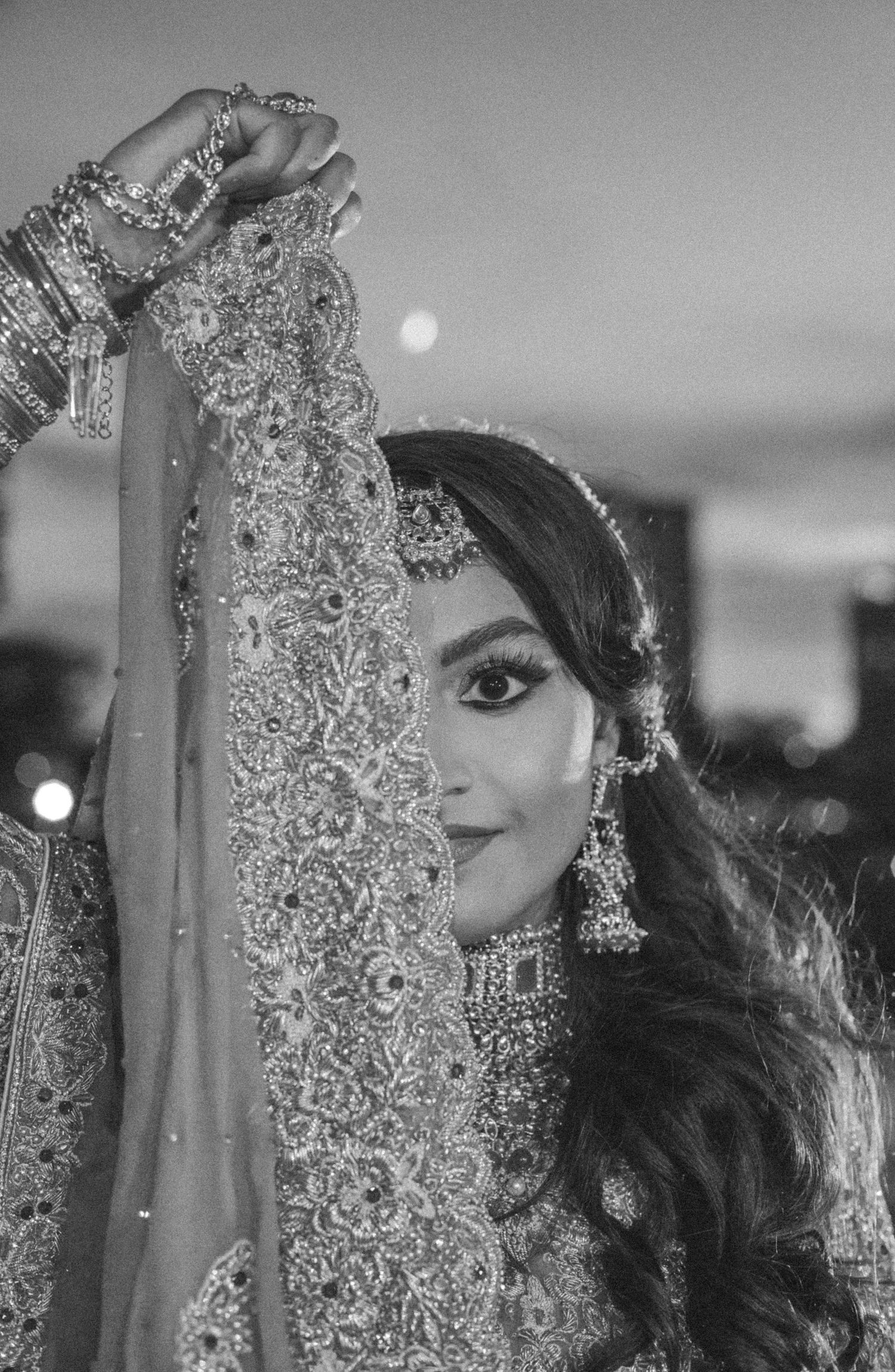 Black and white portrait of a bride peeking from behind a raised embroidered veil with bangles and statement earrings.
