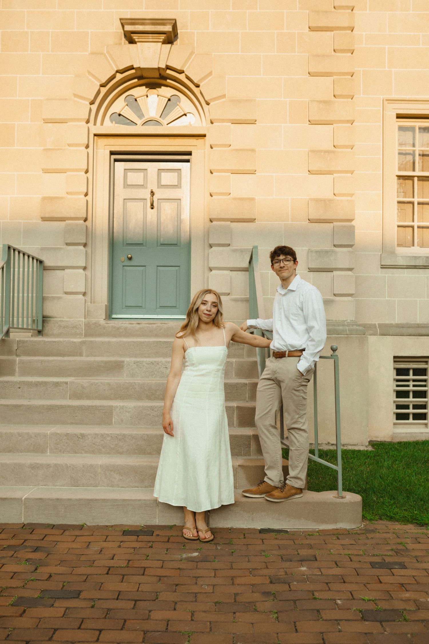 A couple in a white dress and light shirt pose on brick pavers by the green door of a historic stone building at sunset.