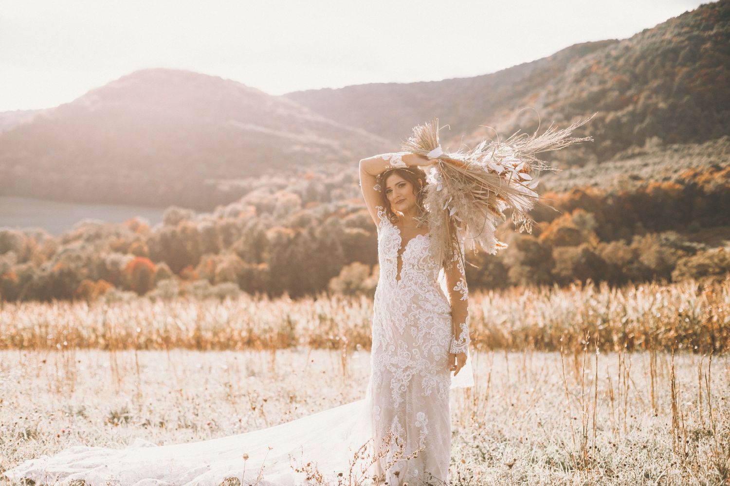 Bride in a lace wedding gown raises her arms and holds a pampas grass bouquet in a golden field with mountains behind her.