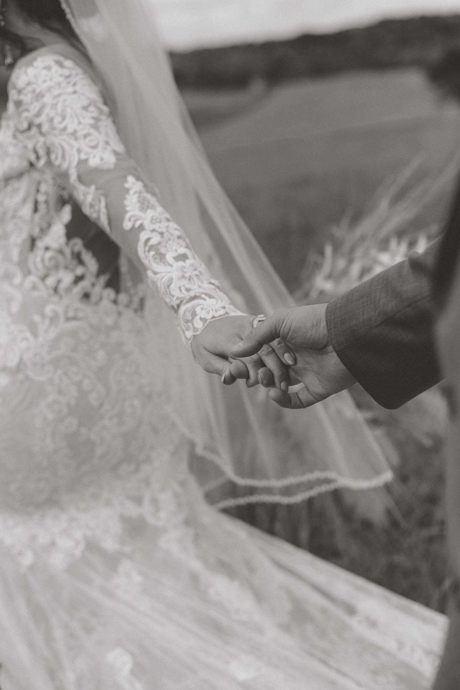 Black and white close-up of a bride in a lace sleeve and veil holding hands with her groom in a grassy field.