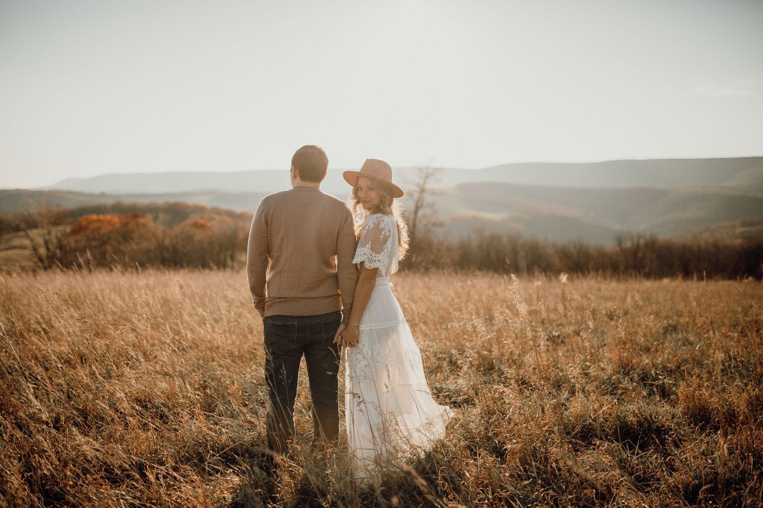 Couple in casual and flowing white outfits stand hand in hand in a golden grass field with distant hills at sunset.