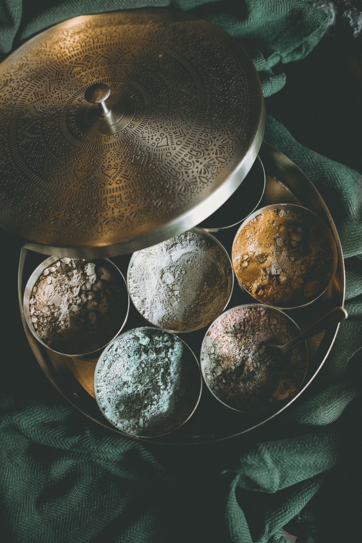 Round engraved brass spice tin holding colorful mineral powders and clays, set on dark green fabric.