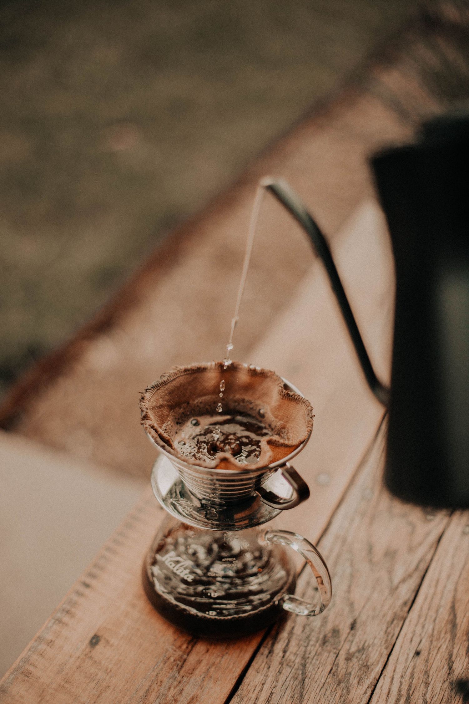 Hot water pouring from a gooseneck kettle into a glass pour-over coffee dripper on a rustic wooden table outdoors.