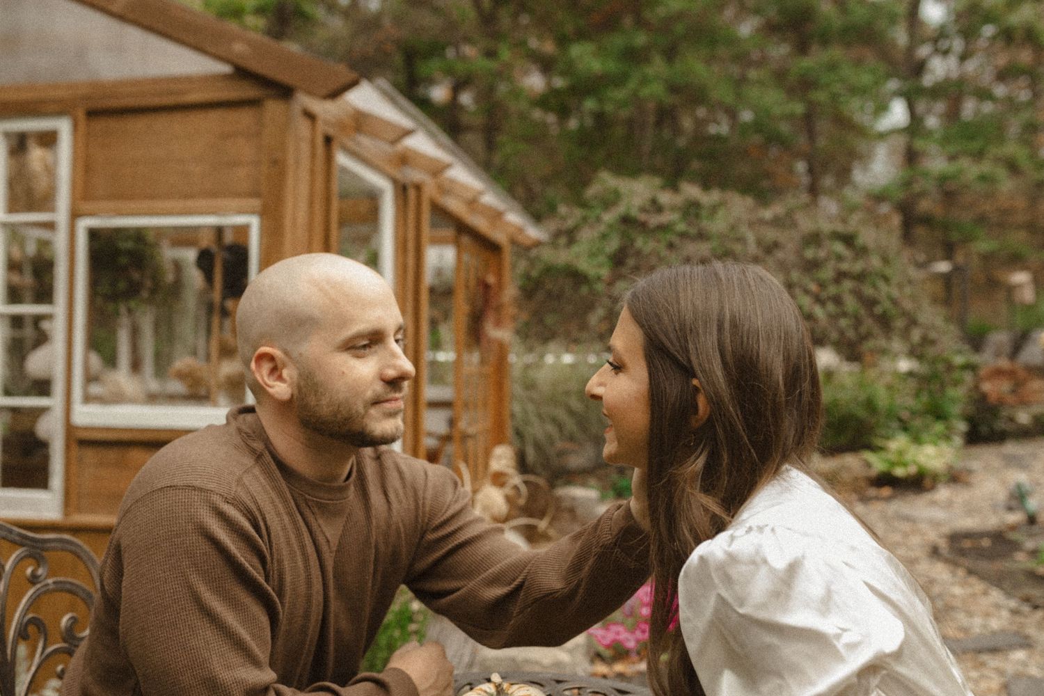 Smiling bald man in a brown sweater gazes at a woman with long hair near a wooden greenhouse in a garden.