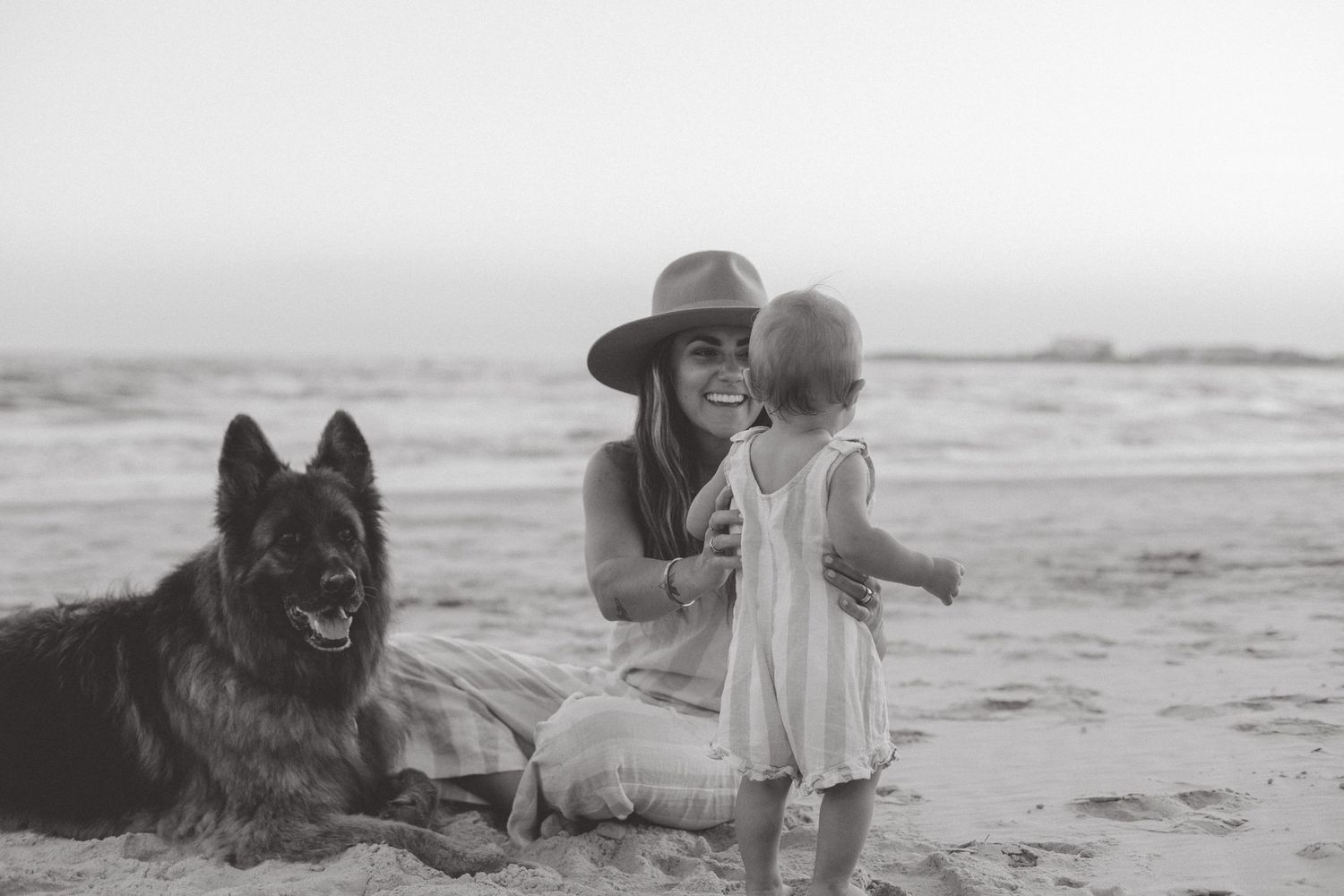 Black and white photo of a mother in a hat and her toddler sitting on the beach beside a large dog.