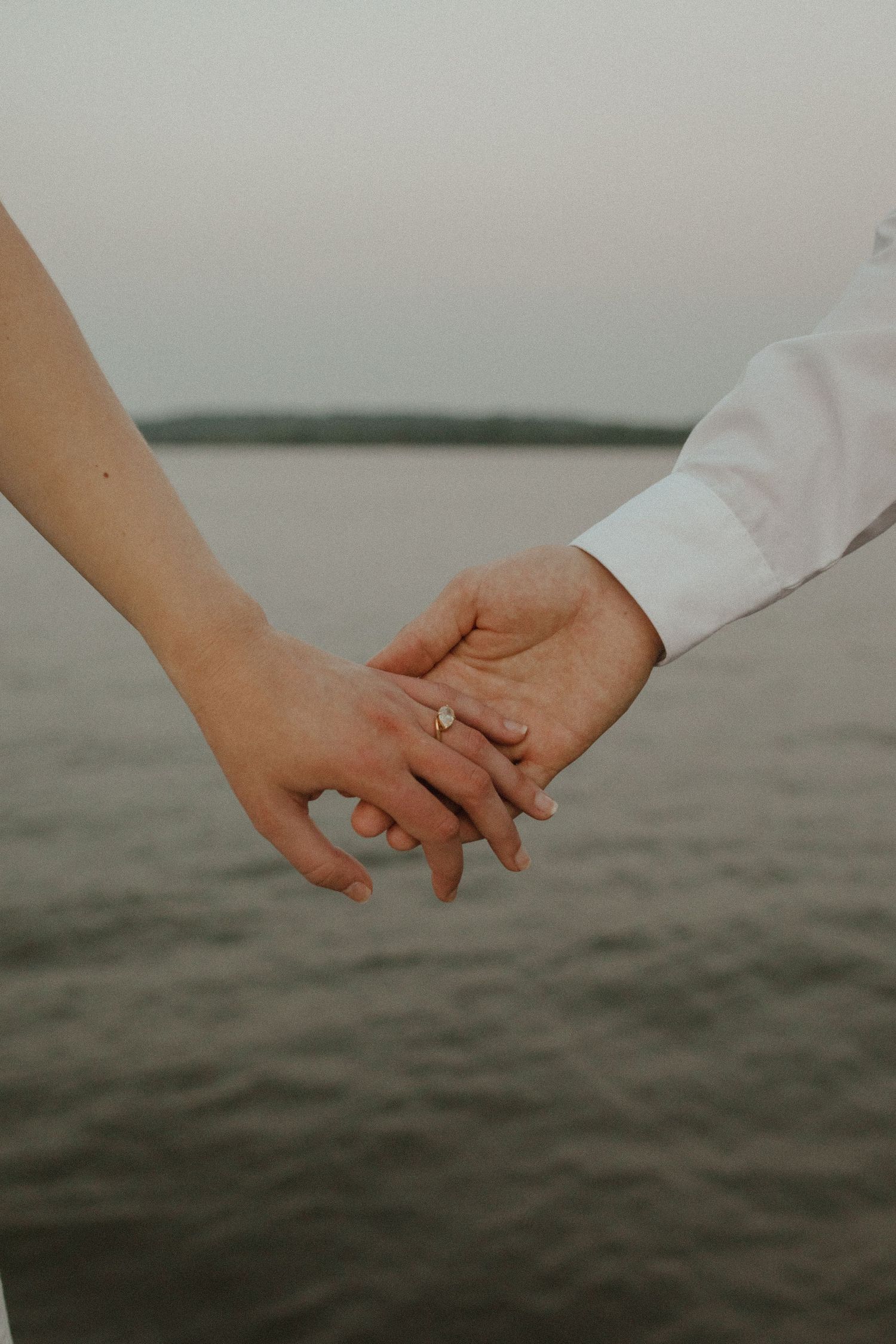 Close-up of a couple holding hands with an engagement ring visible, overlooking a calm river at dusk.