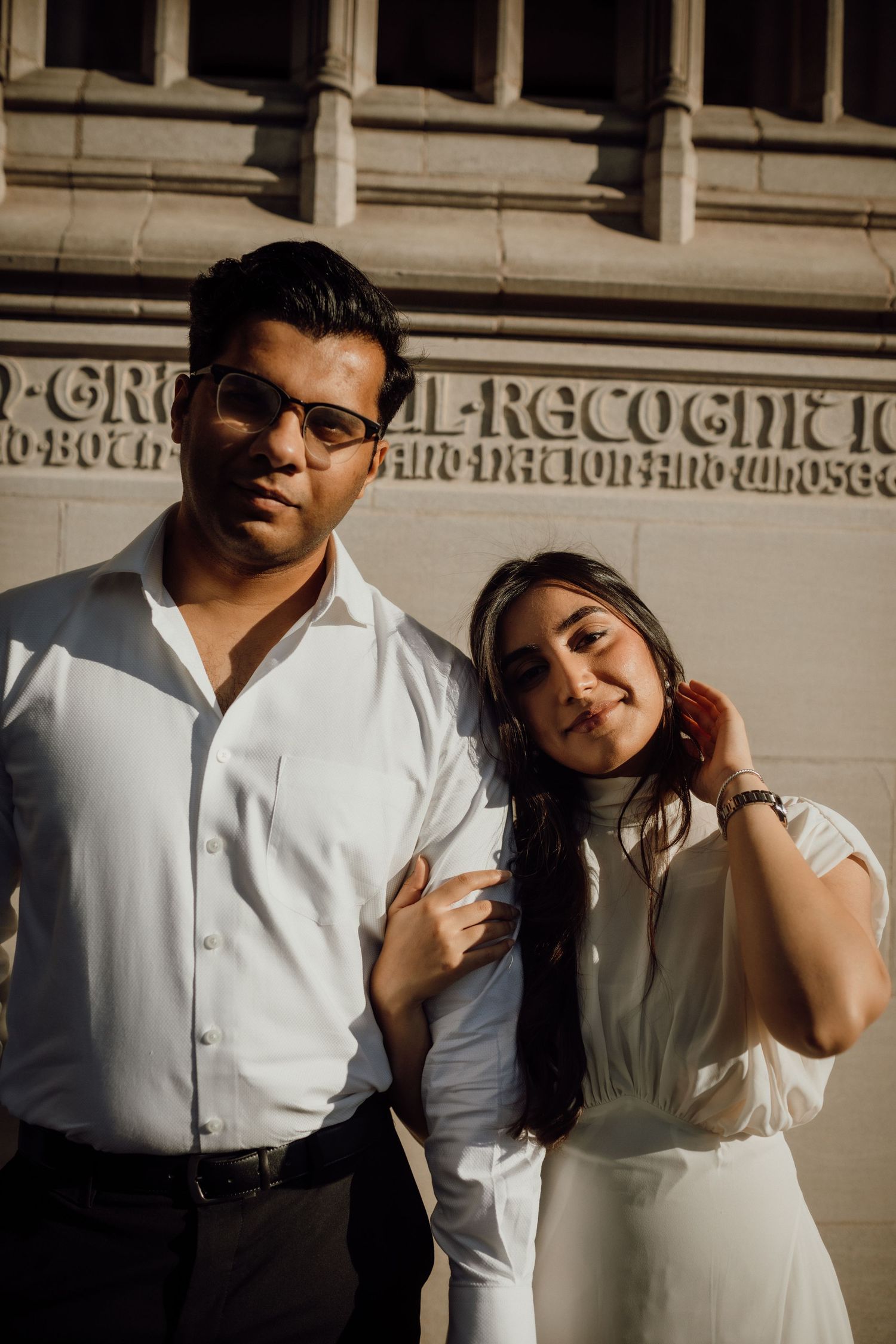 A man with glasses in a white shirt stands beside a smiling woman in white against a sunlit stone building.