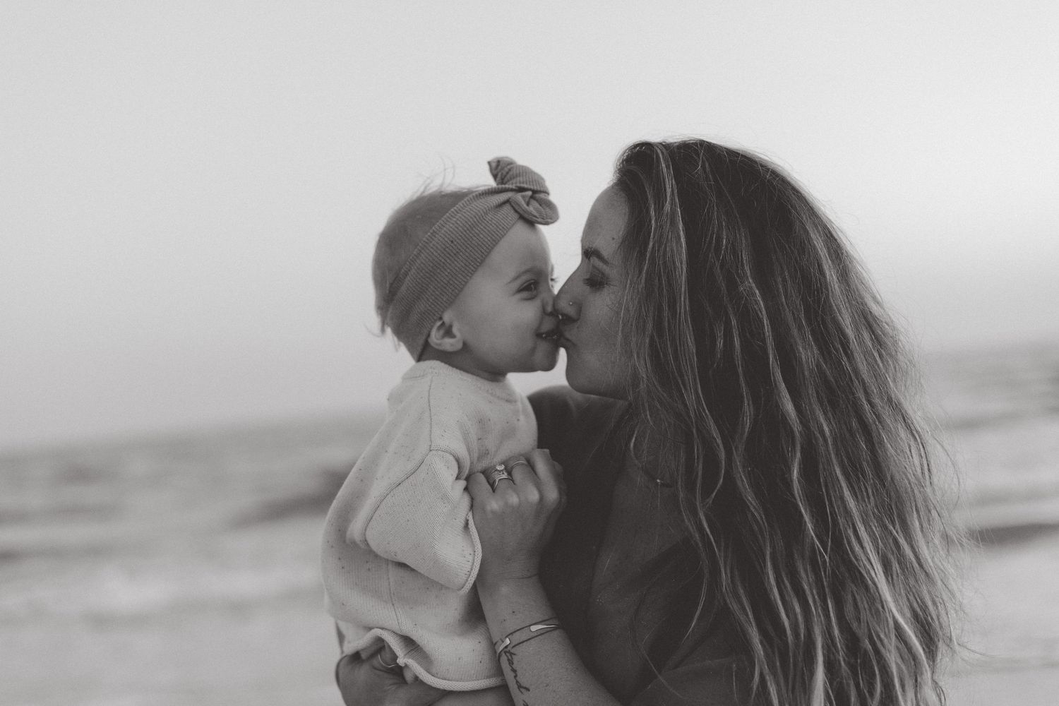 Black and white photo of a mother kissing her baby in a bow headband while standing on a beach.