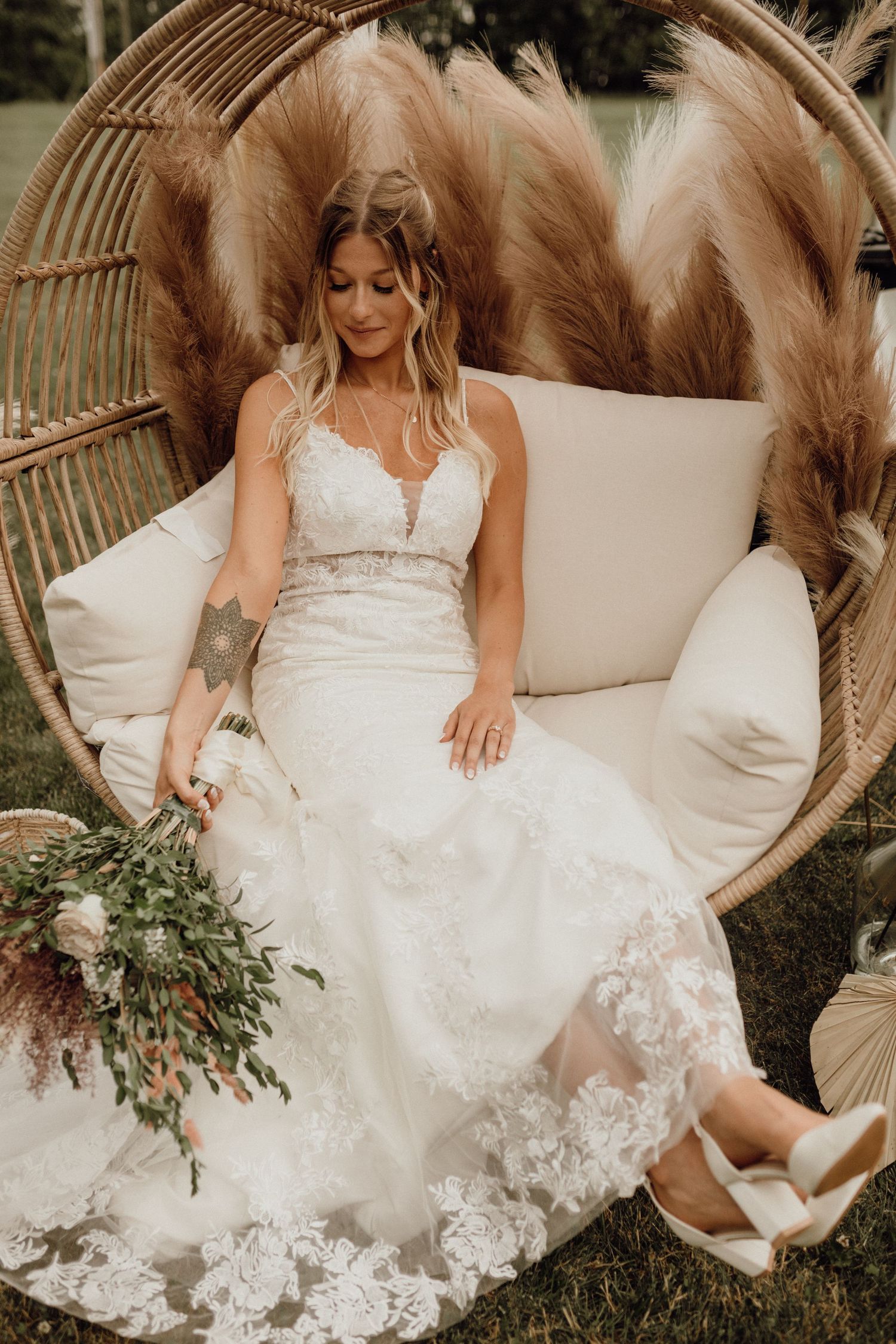 Bride in a lace wedding gown sits in a rattan hanging chair with pampas grass, holding a floral bouquet.