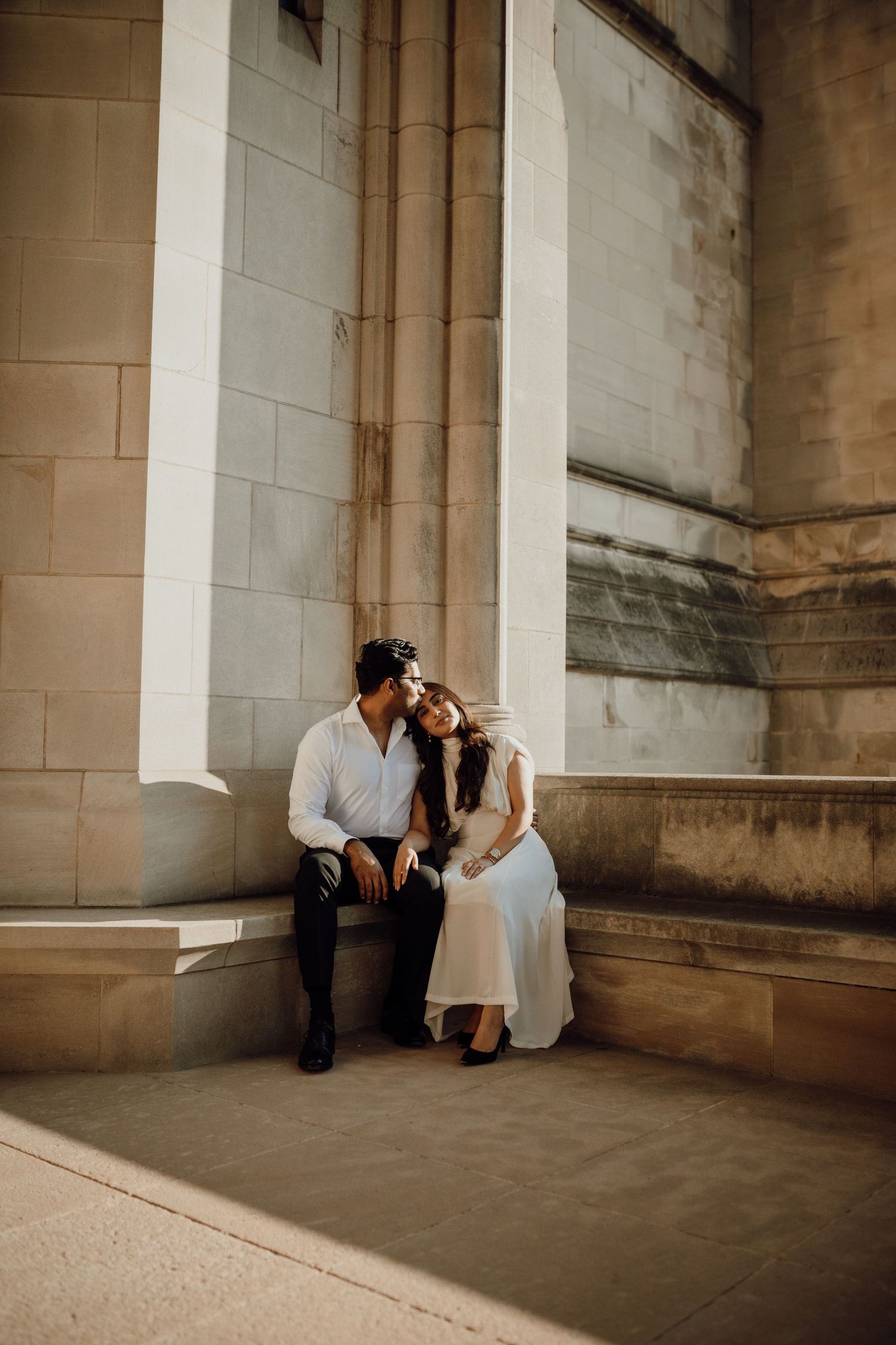 A couple in white and black sit close together on a stone ledge of a sunlit historic stone building.