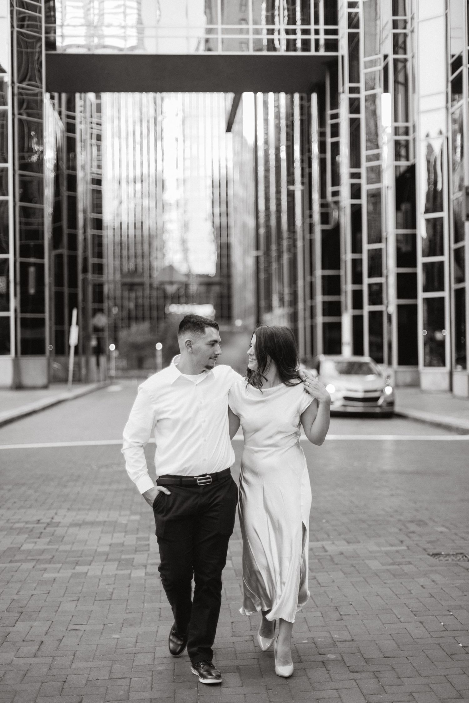 Black and white photo of a couple walking arm in arm down a city street lined with tall glass buildings.