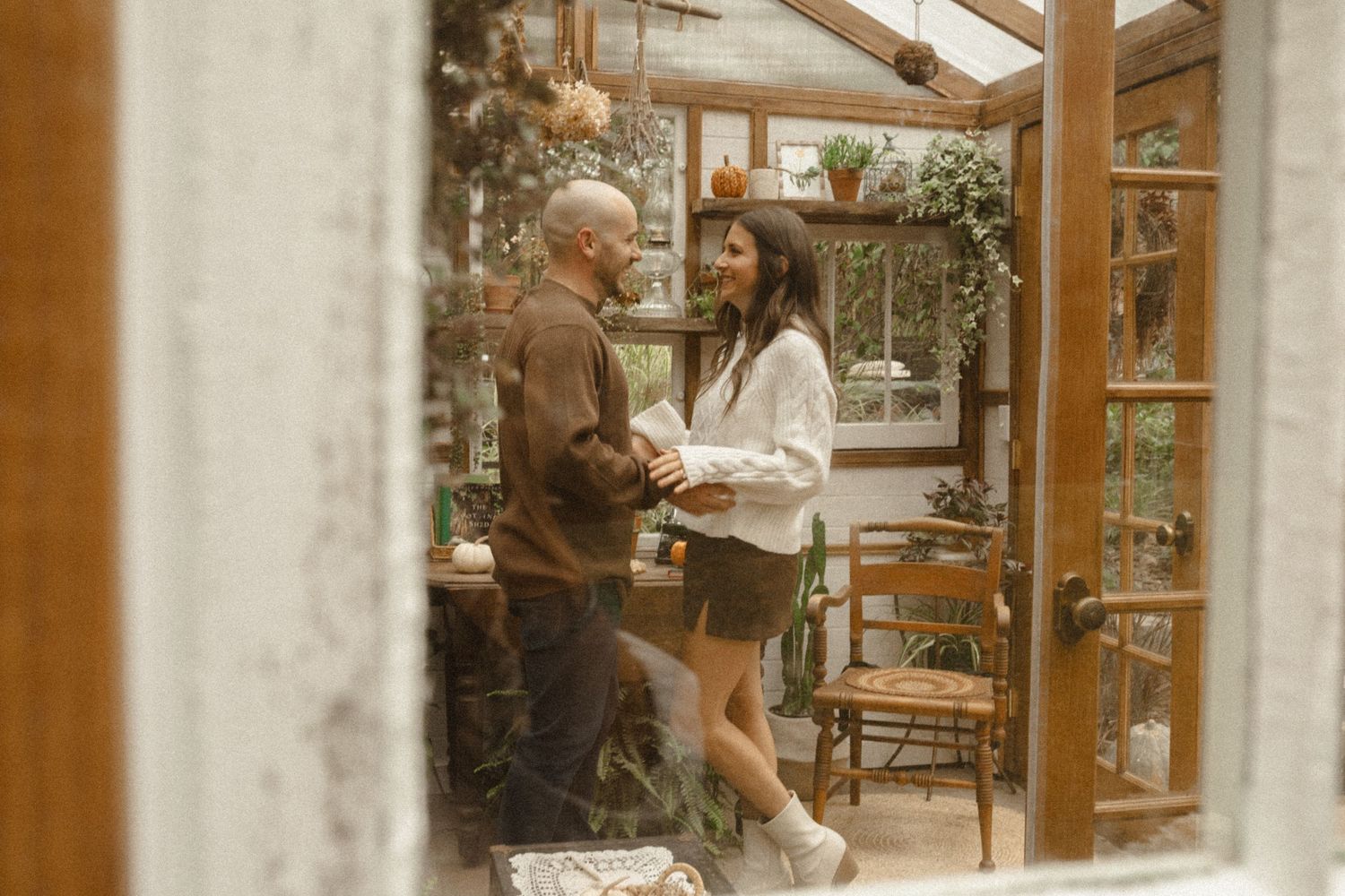 Couple dancing together inside a rustic wooden greenhouse filled with plants, seen through a window frame.