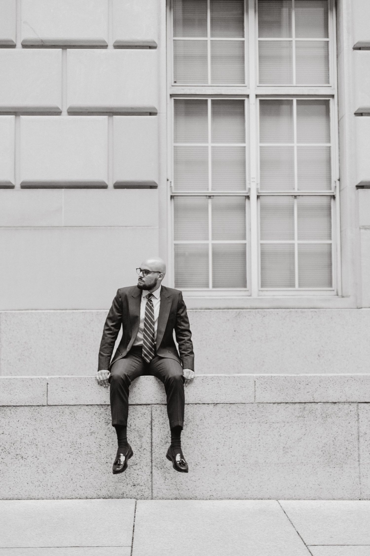 Black and white photo of a bearded man in a suit and glasses sitting on a stone ledge beside a large window.
