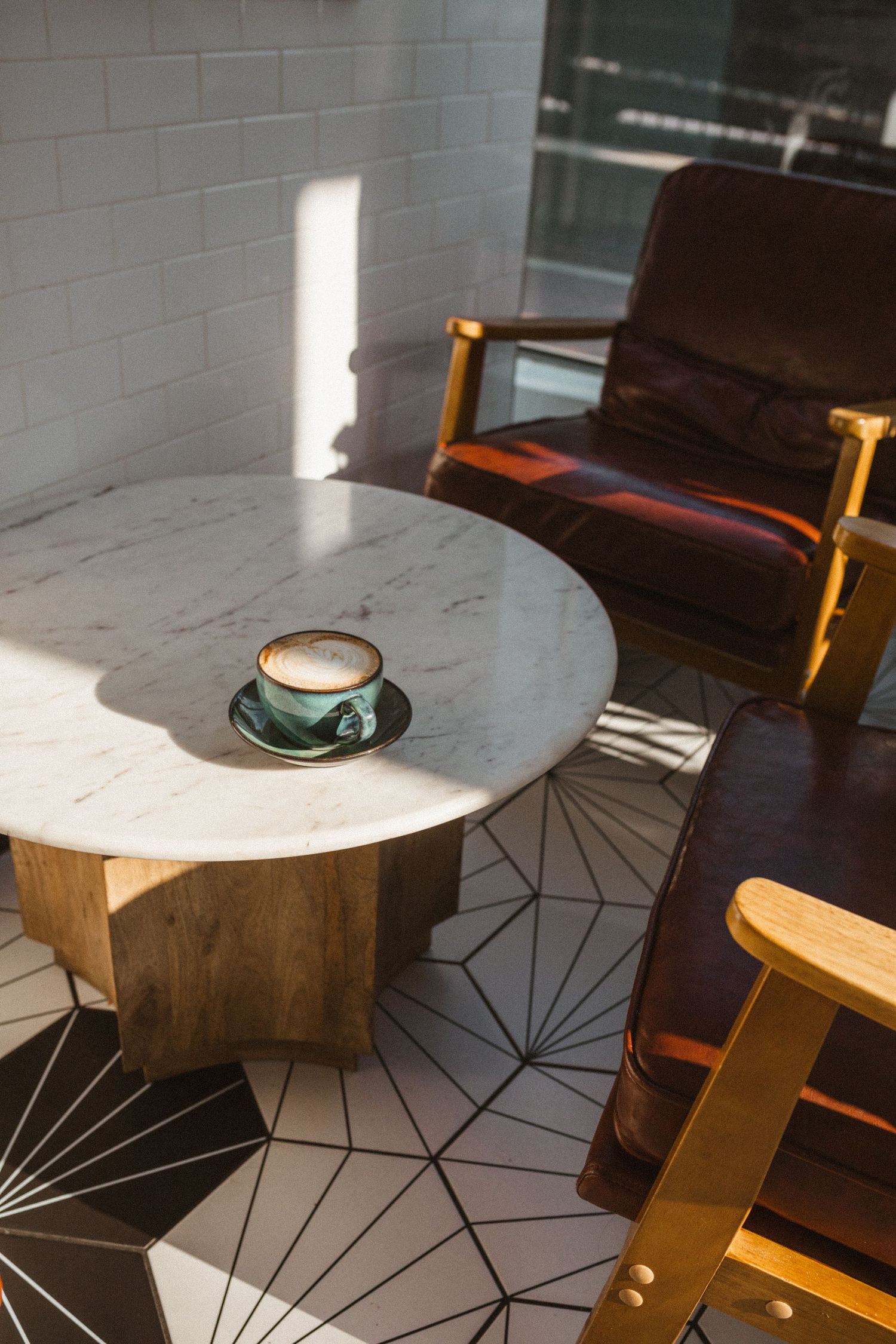 Sunlit marble coffee table with a green cup of latte beside leather armchairs on a geometric tile floor.