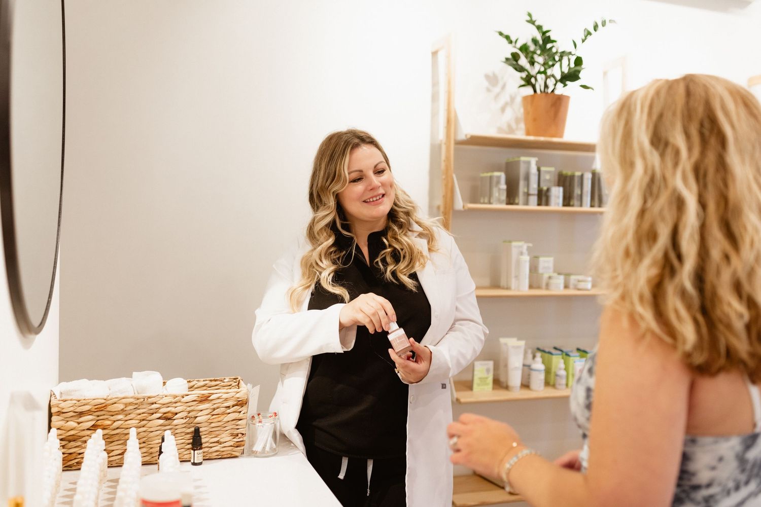 Smiling esthetician in a white coat shows a skincare product to a client in a bright boutique studio.