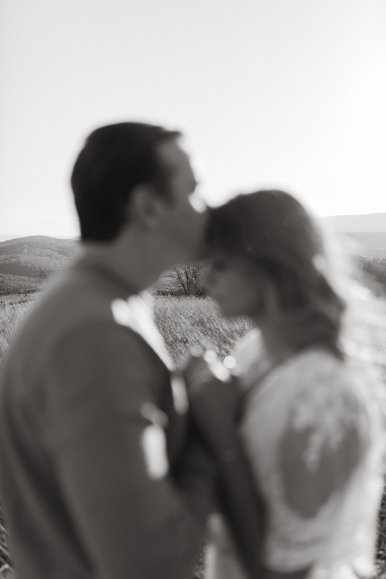 Blurred black and white image of a man kissing his fiancée's forehead with rolling hills in the background.