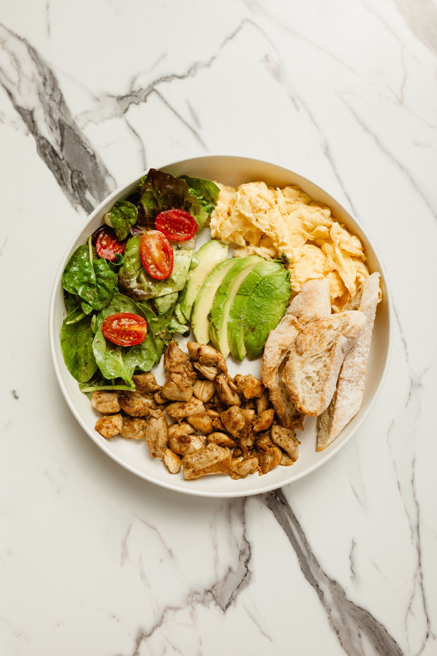 Breakfast bowl with scrambled eggs, sliced avocado, grilled chicken, roasted potatoes, tomatoes and greens on marble.