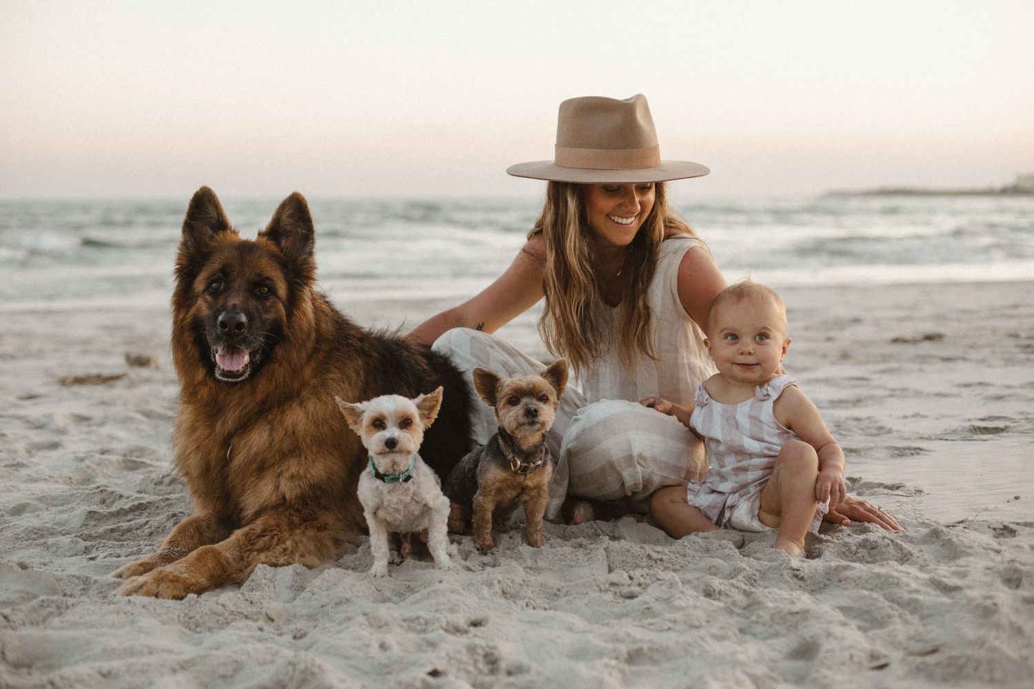 Woman in a straw hat sits on a sandy beach at sunset with a baby, a German Shepherd, and two small dogs.