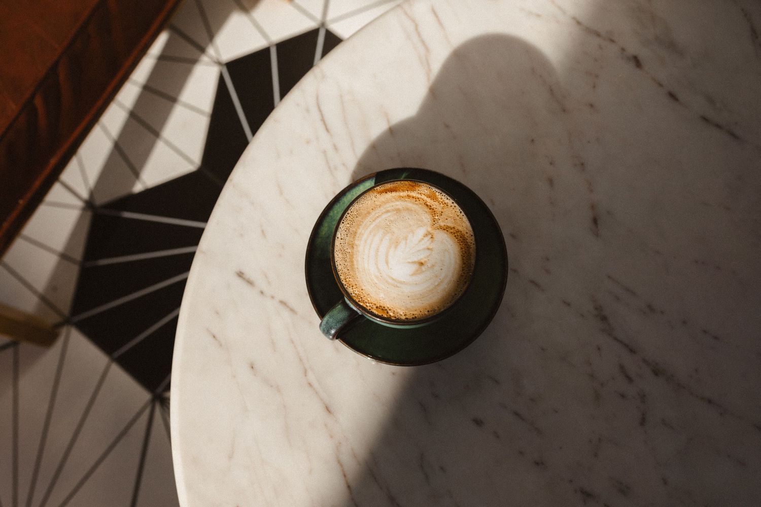 Overhead view of a latte with foam art in a green cup on a white marble café table with geometric tile shadows.