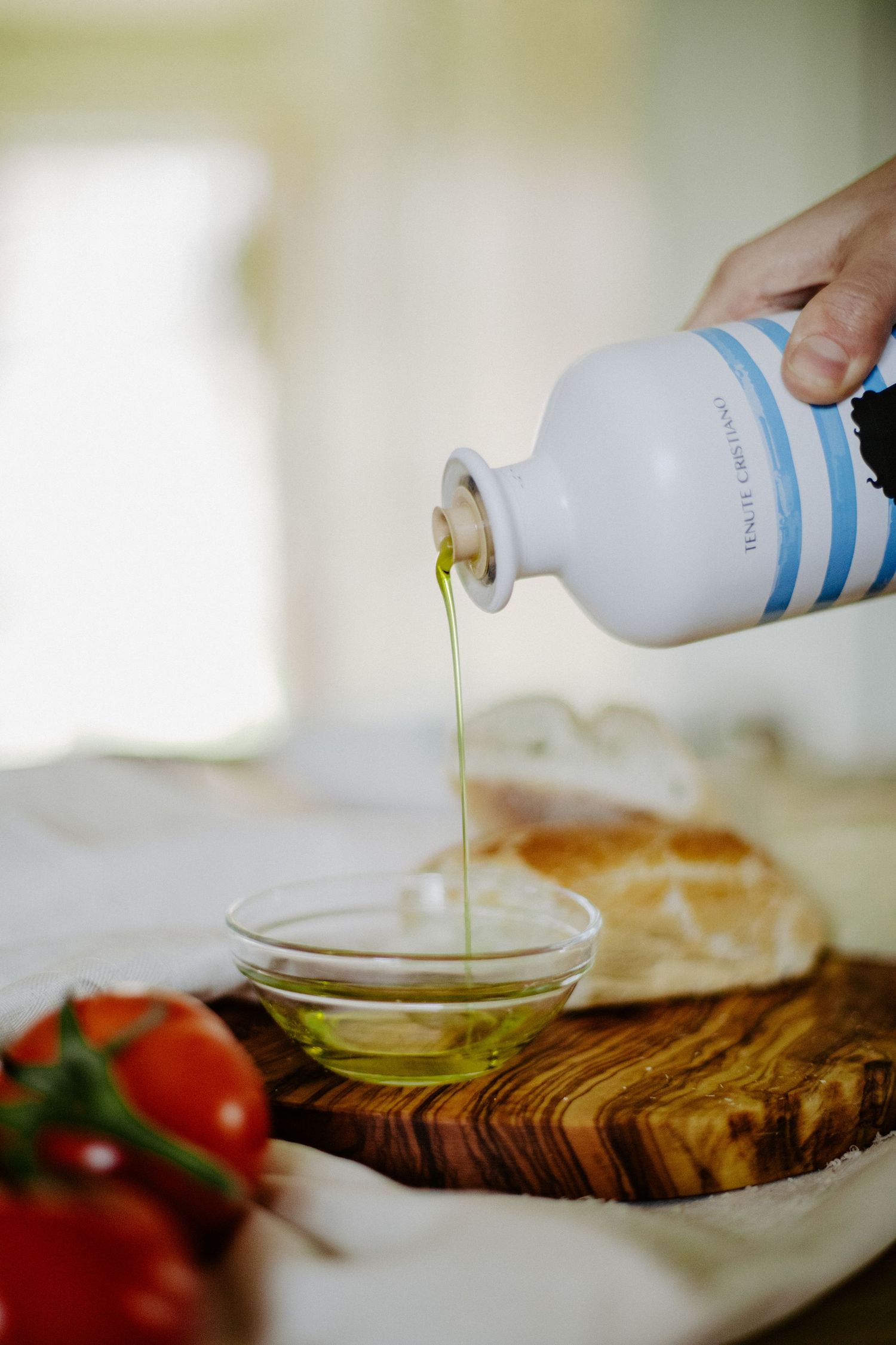 Hand pouring extra virgin olive oil from a ceramic bottle into a glass dish beside bread and tomatoes.
