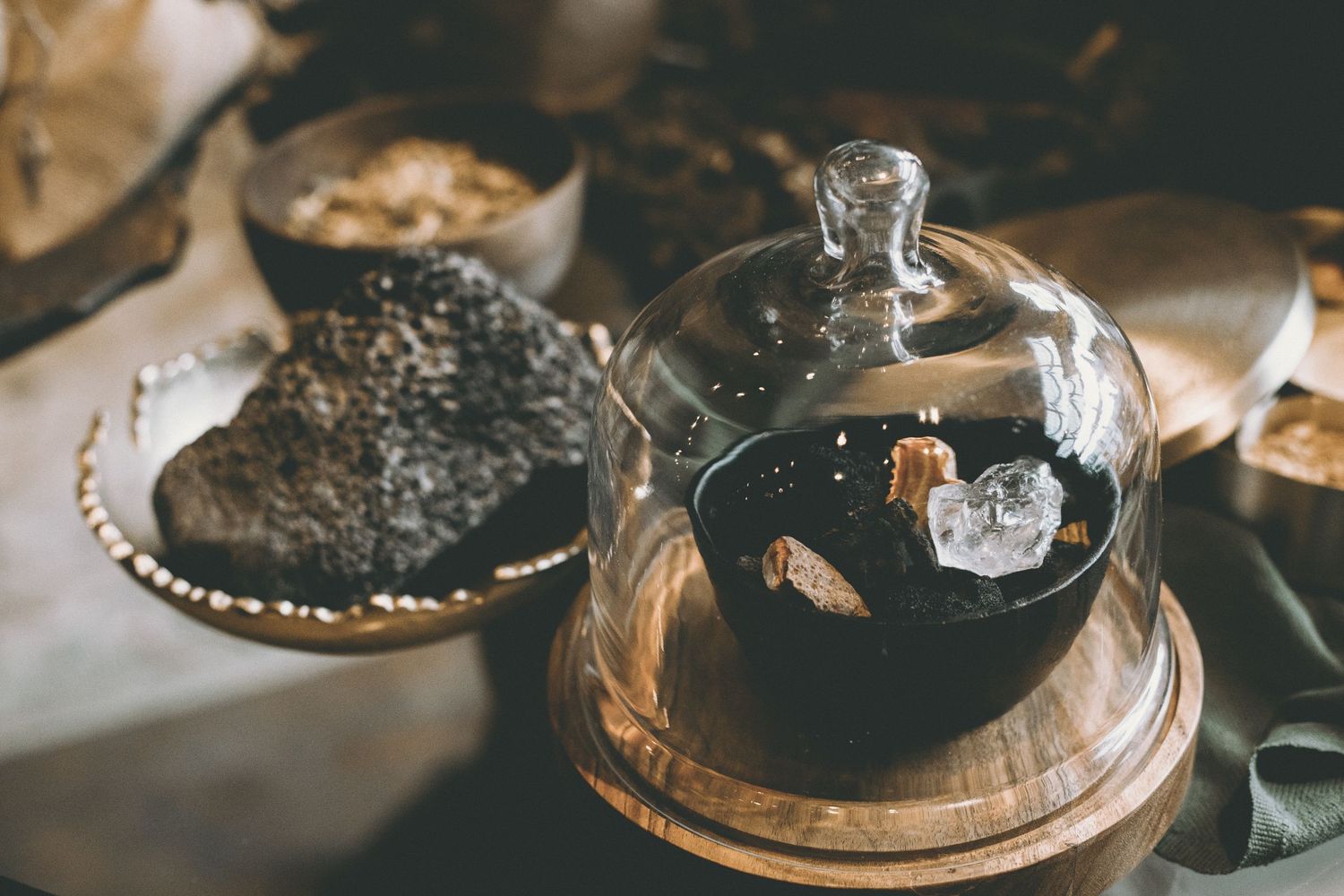 Dark rustic stone on a metal plate beside a glass cloche covering a bowl with crystals on a wooden stand.